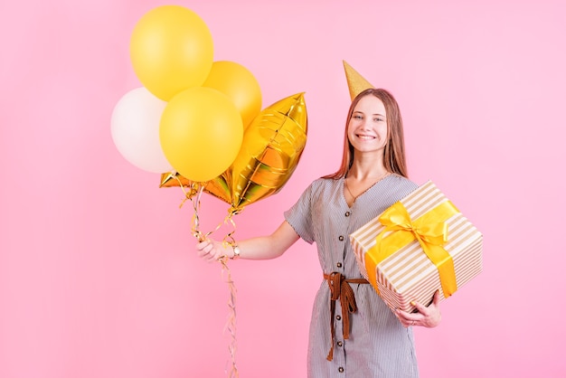 Przyjęcie Urodzinowe. Młoda Kobieta W Urodzinowym Kapeluszu, Trzymając Balony I Duże Pudełko Z Okazji Urodzin Na Różowym Tle Z Miejsca Na Kopię