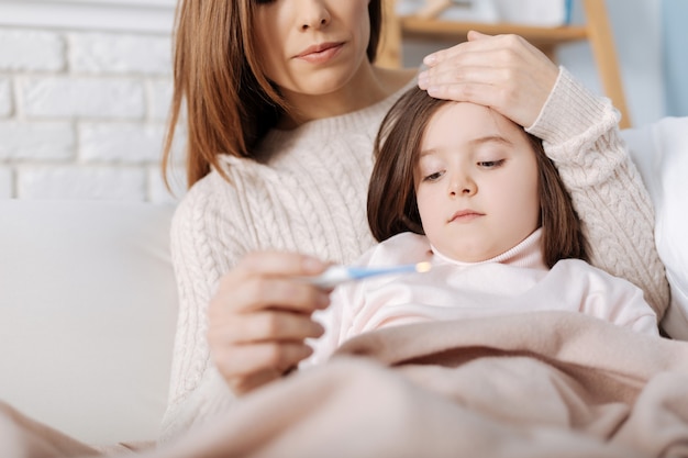 Przyjemna chora dziewczynka leży na sofie, podczas gdy jej troskliwa mama mierzy temperaturę i pomaga w rekonwalescencji