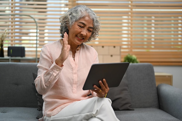 Przyjemna kobieta w średnim wieku sprawdzająca media społecznościowe wykonując rozmowę wideo na cyfrowym tabletie