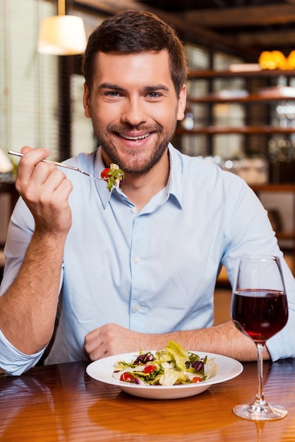 Przyjemny posiłek w restauracji. Przystojny młody mężczyzna je sałatkę i uśmiecha się siedząc w restauracji