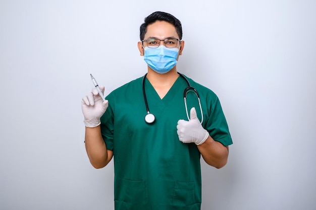 Przyjemny uśmiechnięty azjatycki lekarz mężczyzna w masce medycznej i rękawiczkach pokazuje kciuk w górę trzyma strzykawkę ze szczepionką