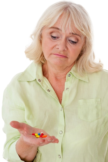 Przyjmowanie tabletek. Przygnębiona starsza kobieta trzymająca pigułki w dłoni i patrząca na nie, gdy jest odizolowana na białym tle