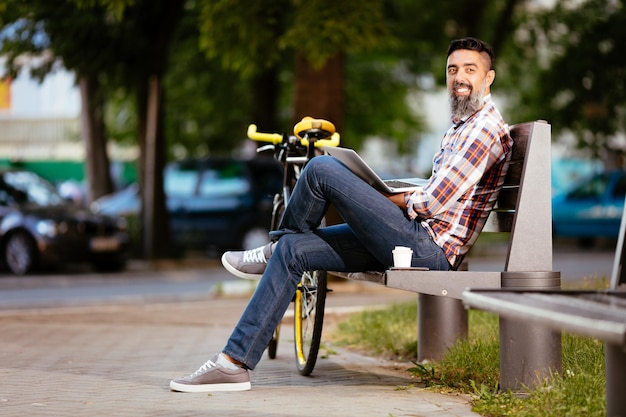 Przypadkowy biznesmen na przerwę na kawę. Siedzi na ławce i pracuje przy laptopie, obok ławki spoczywa rower.