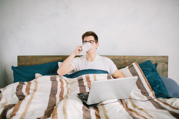 Przypadkowy młody człowiek używa laptop w łóżku w domu i pijący kawę