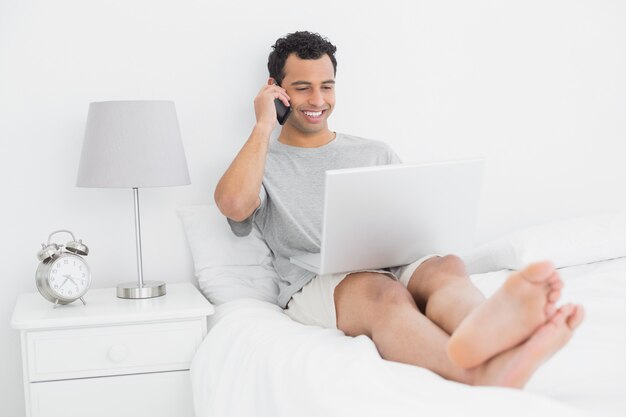 Przypadkowy uśmiechnięty mężczyzna używa telefon komórkowego i laptop w łóżku