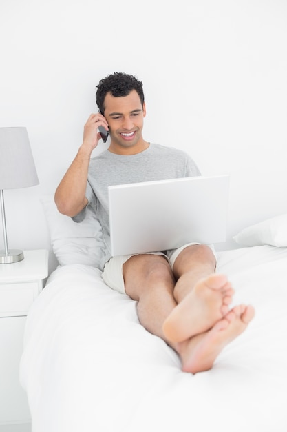 Przypadkowy uśmiechnięty mężczyzna używa telefon komórkowego i laptop w łóżku