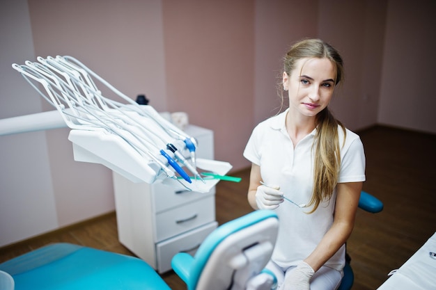 Przystojna Dentystka Pozuje Z Instrumentami Dentystycznymi W Dłoni W Białym Fartuchu W Nowoczesnej, Dobrze Wyposażonej Gabinecie