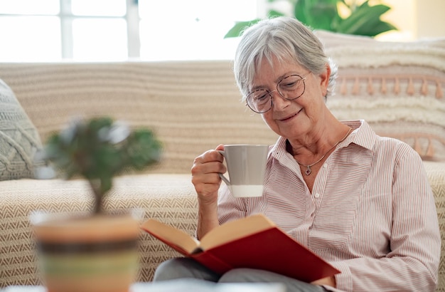 Przystojna kobieta wyższych siedzi na podłodze w domu, czytając książkę, delektując się filiżanką herbaty Uśmiechnięta dojrzała dama zrelaksowana na emeryturze