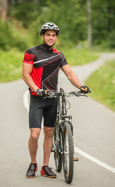 Przystojna mężczyzna pozycja z jego bicyklem na lasowej drodze.