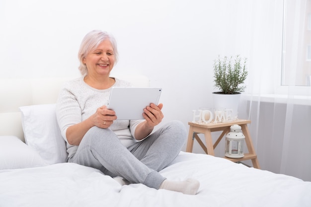 Przystojne starsze kobiety siedzące w łóżku przy użyciu tabletu w domu, mające wideorozmowę z dziećmi.