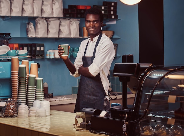 Przystojny African American barista w mundurze trzyma filiżankę kawy, stojąc w swojej kawiarni.