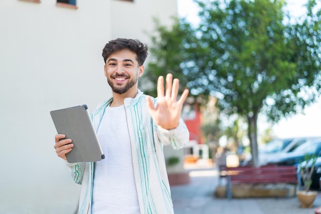 Przystojny Arab trzymający tablet na świeżym powietrzu, salutujący ręką z radosnym wyrazem twarzy