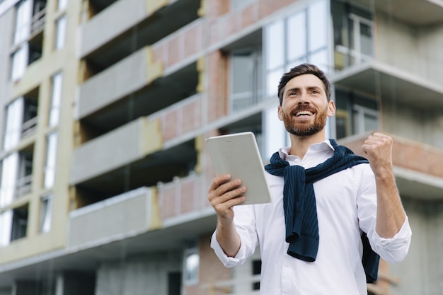 Przystojny architekt, stojąc na placu budowy, wygląda na zadowolonego z ukończonego projektu. Brodaty mężczyzna za pomocą cyfrowego tabletu do pracy.