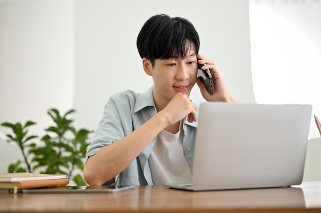 Przystojny Azjata rozmawia z kimś przez telefon, patrząc na ekran swojego laptopa