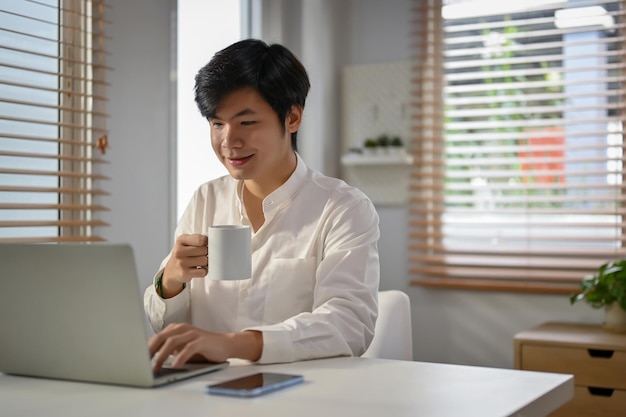 Przystojny azjatycki biznesmen popijający kawę i pracujący nad swoimi zadaniami biznesowymi na laptopie