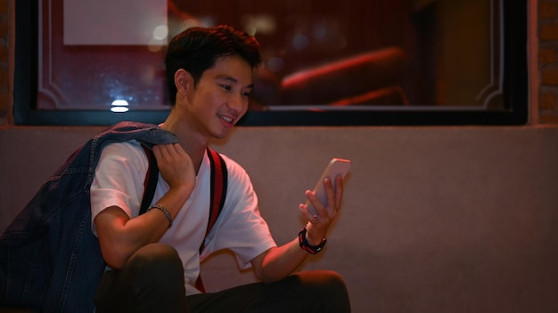 Przystojny azjatycki mężczyzna siedzący w pobliżu gabloty na nocnej ulicy miasta i korzystający z telefonu komórkowego