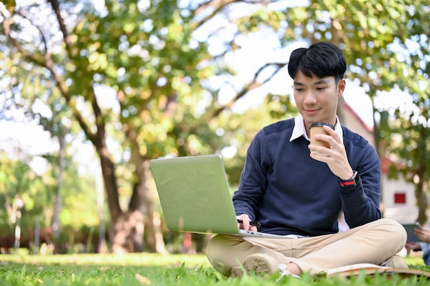 Przystojny azjatycki student college'u za pomocą laptopa popijając kawę siedząc w parku kampusu