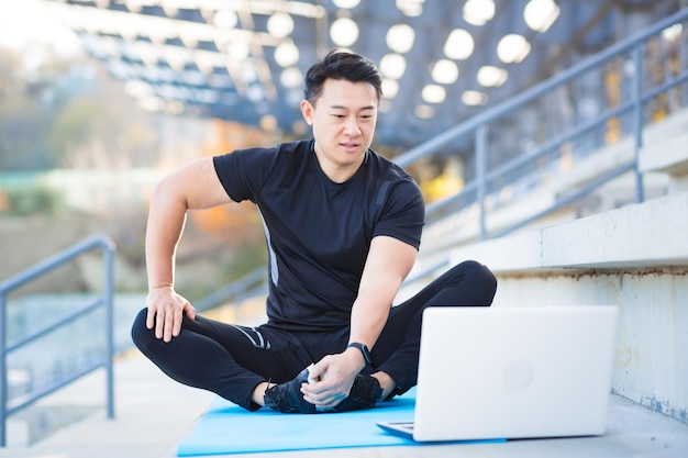 Przystojny azjatycki trener fitness prowadzi zajęcia treningowe online za pomocą kamery internetowej i laptopa, siedząc na zewnątrz na nowoczesnym stadionie Trening Pilates lub rozciąganie Ćwiczenia fizyczne Zapisy Lekcje