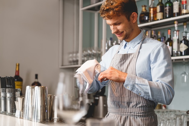 Przystojny barman w fartuchu stojący przy ladzie barowej i czyszczący kieliszek do wina