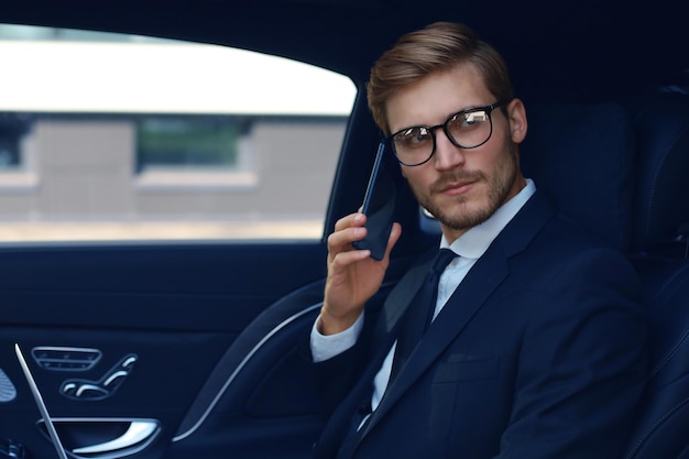 Przystojny biznesmen rozmawia z telefonem, siedząc z laptopem na tylnym siedzeniu samochodu