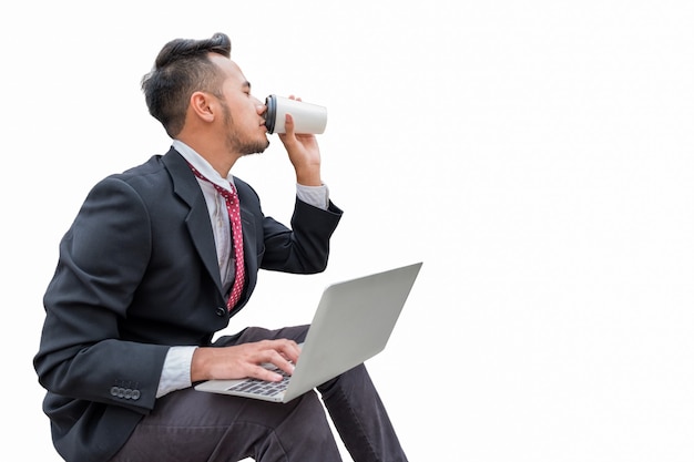 Przystojny biznesmen siedzi i pije kawę za pomocą laptopa, na białym tle