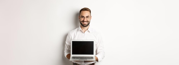 Przystojny biznesmen w okularach pokazujący ekran laptopa i uśmiechnięty szczęśliwy stojący nad białym backgrou
