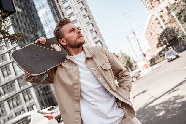 Przystojny blond młody człowiek deskorolkarz trzymając longboard na ramieniu patrząc poważnie na odległość na tle miejskiego budynku. Nosi swobodną beżową koszulę dżinsową. Wypoczynek na świeżym powietrzu. Sport ekstremalny.