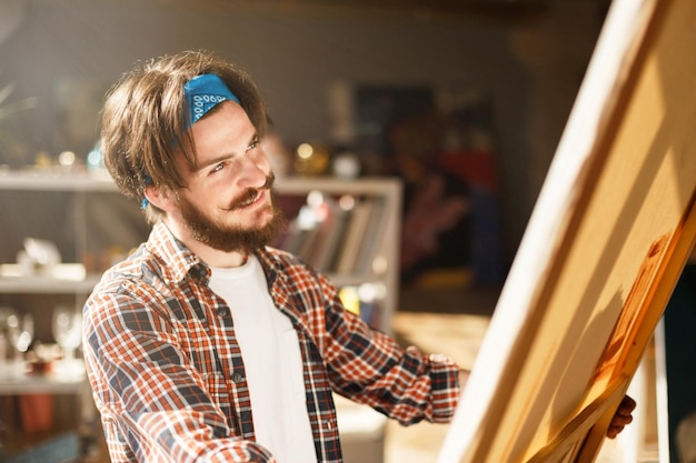 Przystojny, brodaty, czarnowłosy artysta hipster w niebieskiej chustce, patrzący na płótno w jasnym, współczesnym studio