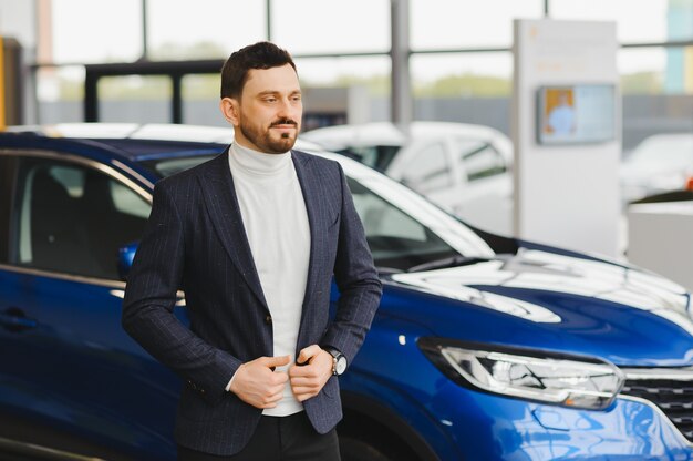 Przystojny brodaty kupiec w codziennym noszeniu w salonie, facet patrzy na kamerę, stojąc w pobliżu samochodu z rozłożonymi rękami