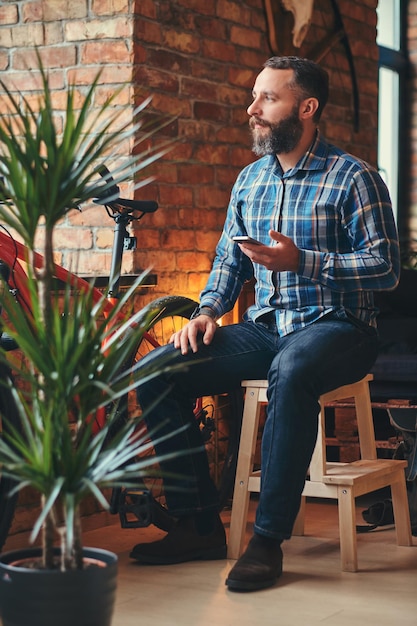 Przystojny brodaty mężczyzna hipster w niebieskiej bluzie z polaru i dżinsach trzyma smartfon, siedząc na drewnianym stołku w studio z wnętrzem na poddaszu.