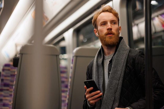 Przystojny brodaty mężczyzna siedzący w metrze ze smartfonem słuchającym muzyki przez słuchawki bezprzewodowe