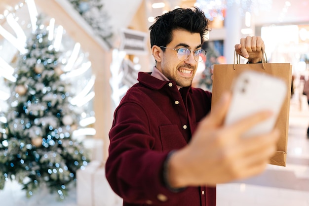 Przystojny brodaty młody mężczyzna patrzący na kamerę robiący selfie przez telefon pozowanie z papierowymi torbami z prezentami stojącymi w holu centrum handlowego świętuj