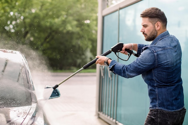 Przystojny brodaty młody mężczyzna w dżinsowej koszuli myje swój nowy samochód ręcznie wężem wysokociśnieniowym w serwisie samoobsługowym na świeżym powietrzu Koncepcja mycia samochodu