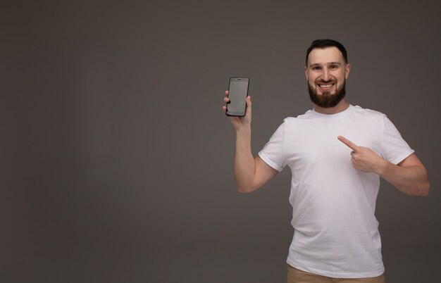 Przystojny brodaty model pozuje z telefonem w rękach nad szarym tłem.