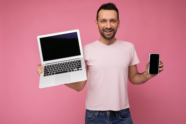 Przystojny Brunet Mężczyzna Trzyma Laptopa I Telefon Komórkowy, Patrząc Na Kamerę W Koszulce Na Białym Tle