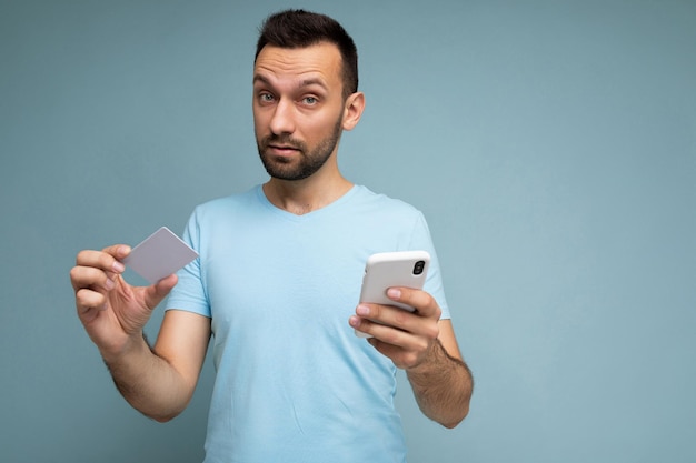 Przystojny brunet mężczyzna ubrany w codzienne ubrania na białym tle na niebieskim tle ściany z miejscem na kopię trzymającą i używającą telefonu komórkowego i plastikowej karty kredytowej dokonującej płatności