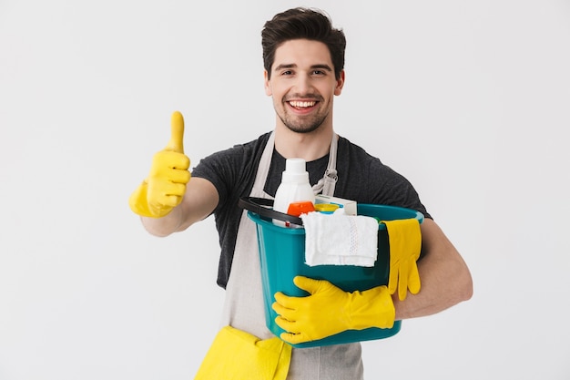 Przystojny Brunetki Houseman Ubrany W Fartuch Stojący Na Białym Tle Nad Białym, Trzymający Wiadro Pełne Detergentów