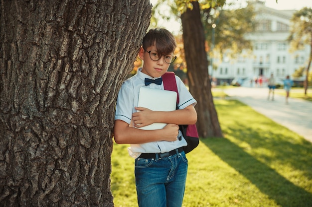 Przystojny chłopiec w wieku szkolnym stoi przy drzewie i trzyma tablet. Uczeń jest ubrany w dżinsy i bez koszuli z muszką.