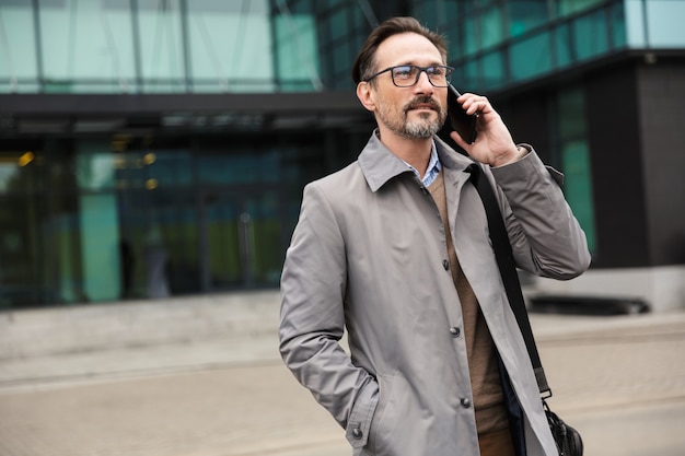przystojny dojrzały biznesmen w okularach rozmawia przez telefon komórkowy, stojąc w pobliżu biurowca w obszarze miejskim