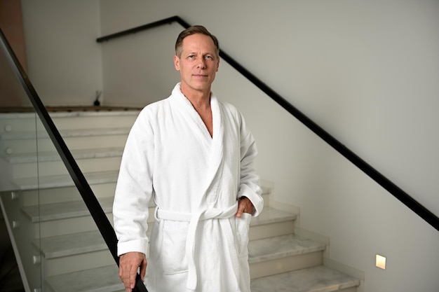 Przystojny dojrzały Europejczyk w białym szlafroku, patrzący pewnie w kamerę, stojąc przy schodach w strefie wypoczynkowej spa wellness, relaksując się i otrzymując męskie zabiegi upiększające
