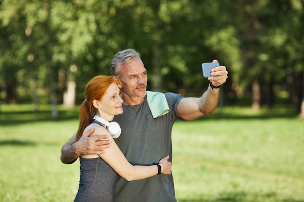 Przystojny dojrzały sportowiec z ręcznikiem na ramieniu obejmujący żonę i wziąć z nią selfie po treningu w parku
