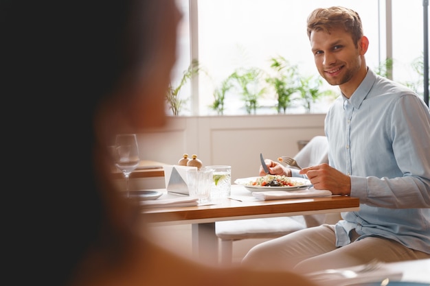 Przystojny dżentelmen patrzący na młodą kobietę i uśmiechający się siedząc przy stole z jedzeniem w restauracji