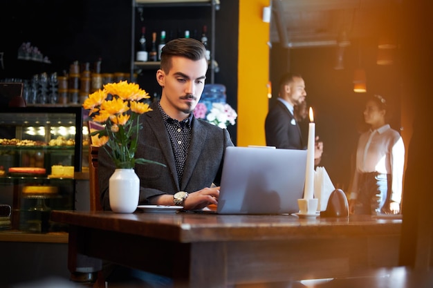 Przystojny freelancer mężczyzna ze stylową brodą i włosami, ubrany w czarny garnitur, pracuje na laptopie, siedząc w kawiarni.