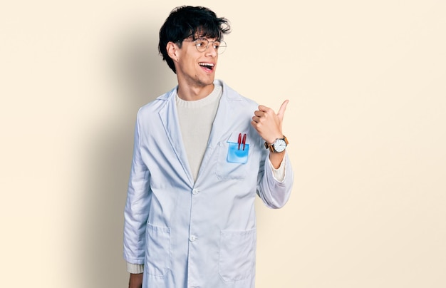 Przystojny hipster młody człowiek ubrany w mundur lekarza wskazujący kciukiem na bok uśmiechnięty zadowolony z otwartymi ustami