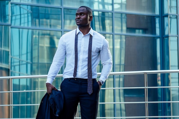 Przystojny i stylowy pan młody Afroamerykanin w modnej czarnej marynarce i białej koszuli z kołnierzykiem z eleganckim krawatem pozuje na tle szklanego pejzażu biurowego Manhattanu