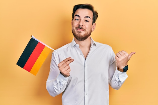 Przystojny kaukaski mężczyzna z brodą trzymający flagę niemiec wskazujący kciukiem na bok uśmiechnięty zadowolony z otwartymi ustami