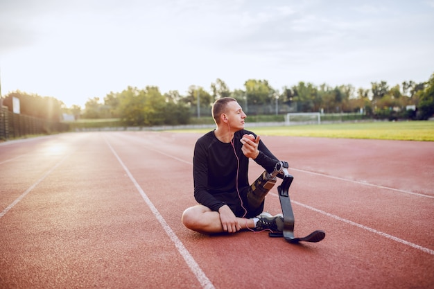 Przystojny kaukaski sportowy niepełnosprawny młody człowiek w odzieży sportowej ze sztuczną nogą, siedzący na torze wyścigowym, słuchając muzyki i jedząc jabłko.