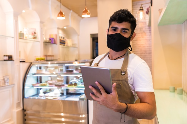 Zdjęcie przystojny kelner w białej koszulce pracujący z tabletem stojący w kawiarni ma na sobie maskę