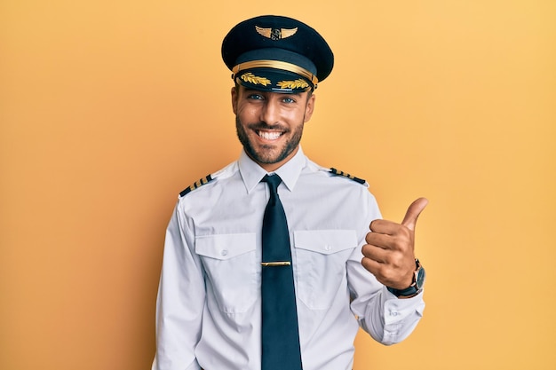 Przystojny latynos ubrany w mundur pilota samolotu, znak sukcesu, wykonujący pozytywny gest z kciukami w górę, uśmiechnięty i szczęśliwy, wesoły wyraz twarzy i gest zwycięzcy