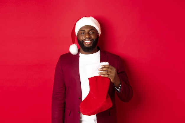 Przystojny mężczyzna afroamerykanin świętuje ferie zimowe, trzymając skarpetę świąteczną i uśmiechnięty, ubrany w santa hat, stojąc na czerwonym tle.